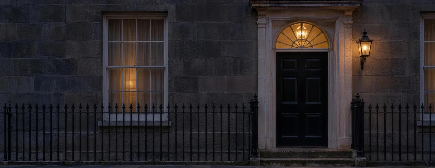 A black Georgian front door in a pale stone surround at dusk, with a lit fanlight above it and a lantern beside it.